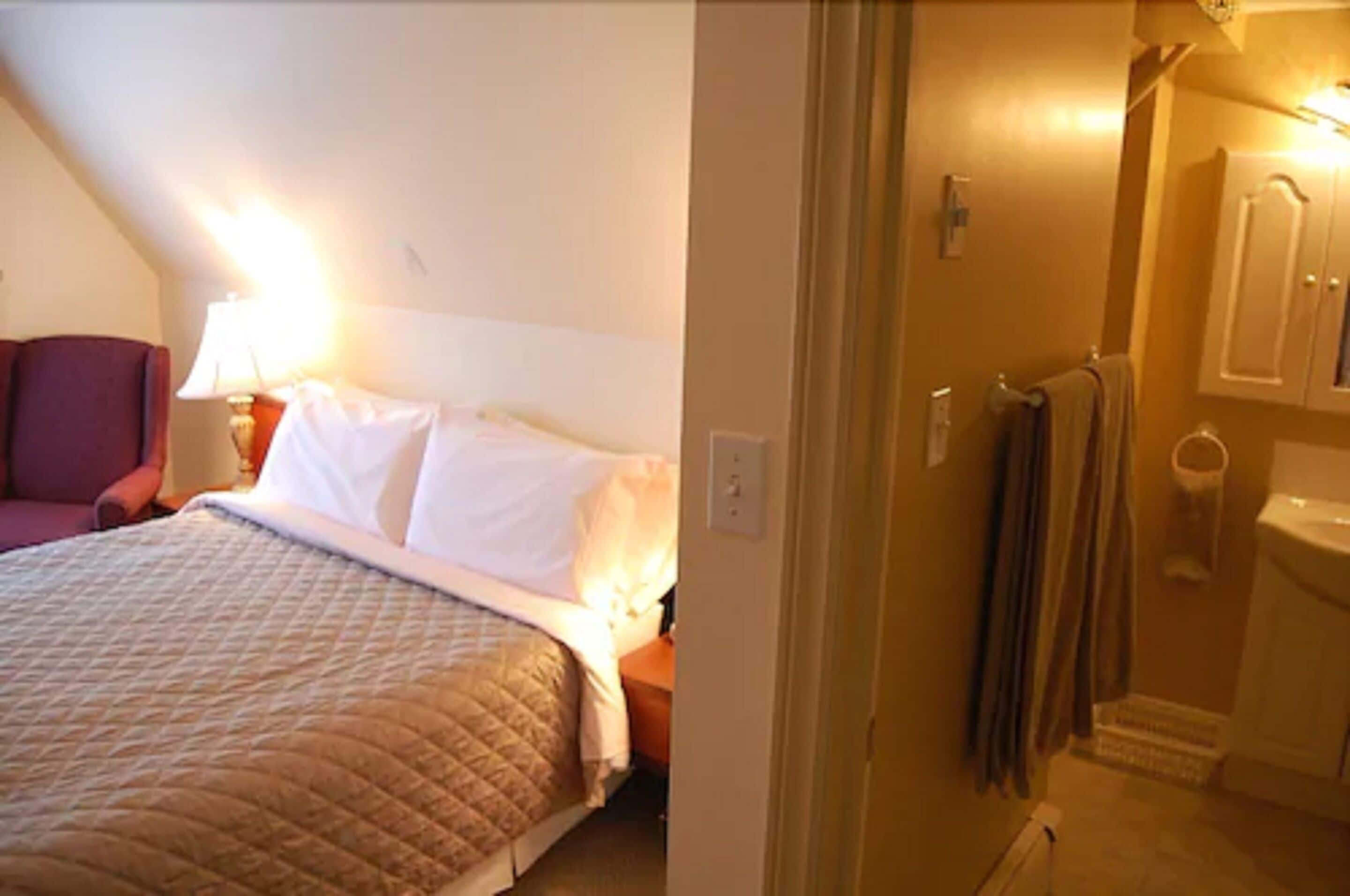 A bedroom with a bed, a nightstand, a lamp, and a view of a bathroom with a toilet and towel rack.