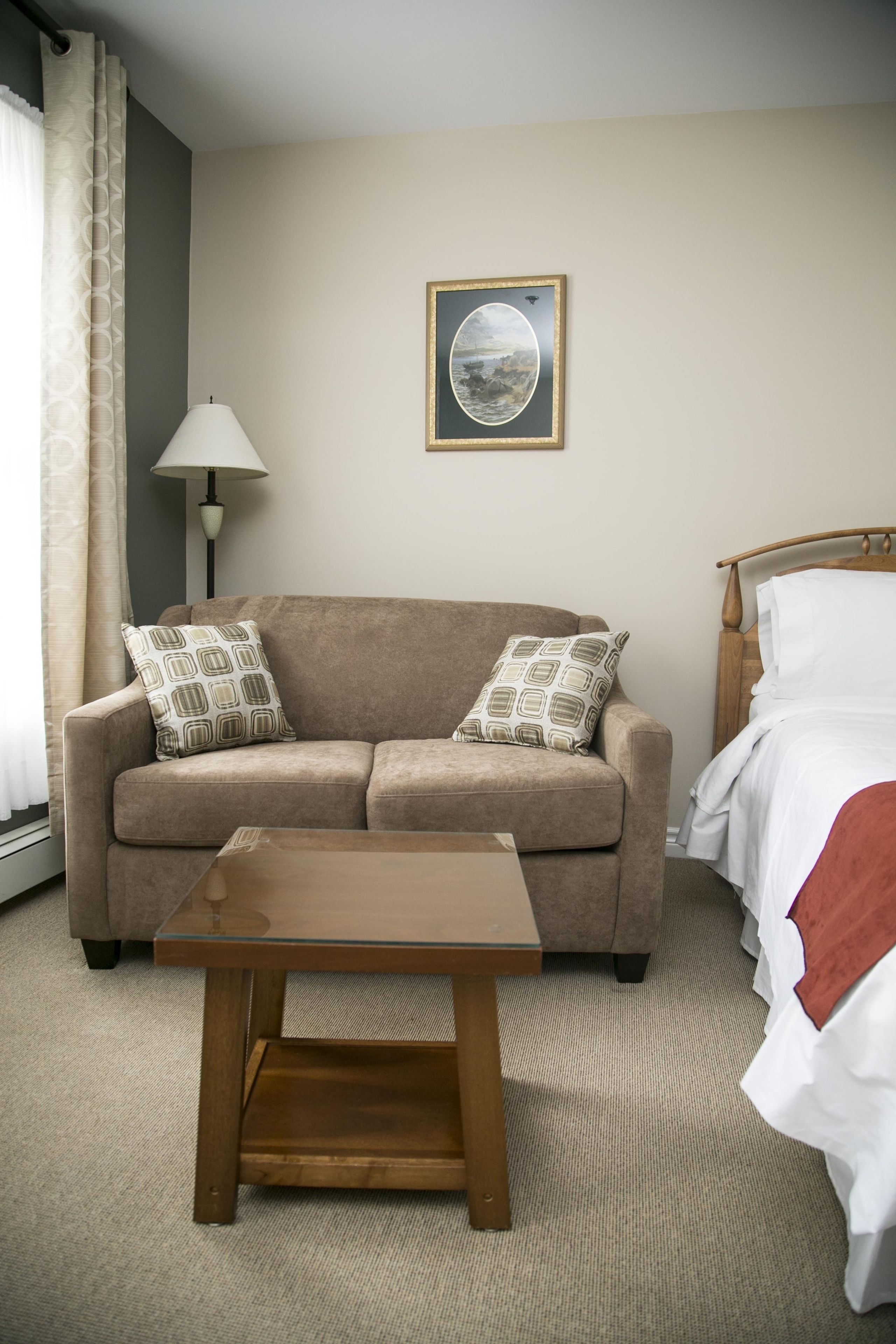 A hotel room with a beige sofa, two patterned pillows, a wooden coffee table, a lamp, a framed picture on the wall, and a bed with white bedding.