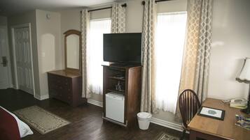Habitació amb televisió sobre un suport de fusta, nevera petita, una cadira, taula amb una làmpada i finestra amb cortines.