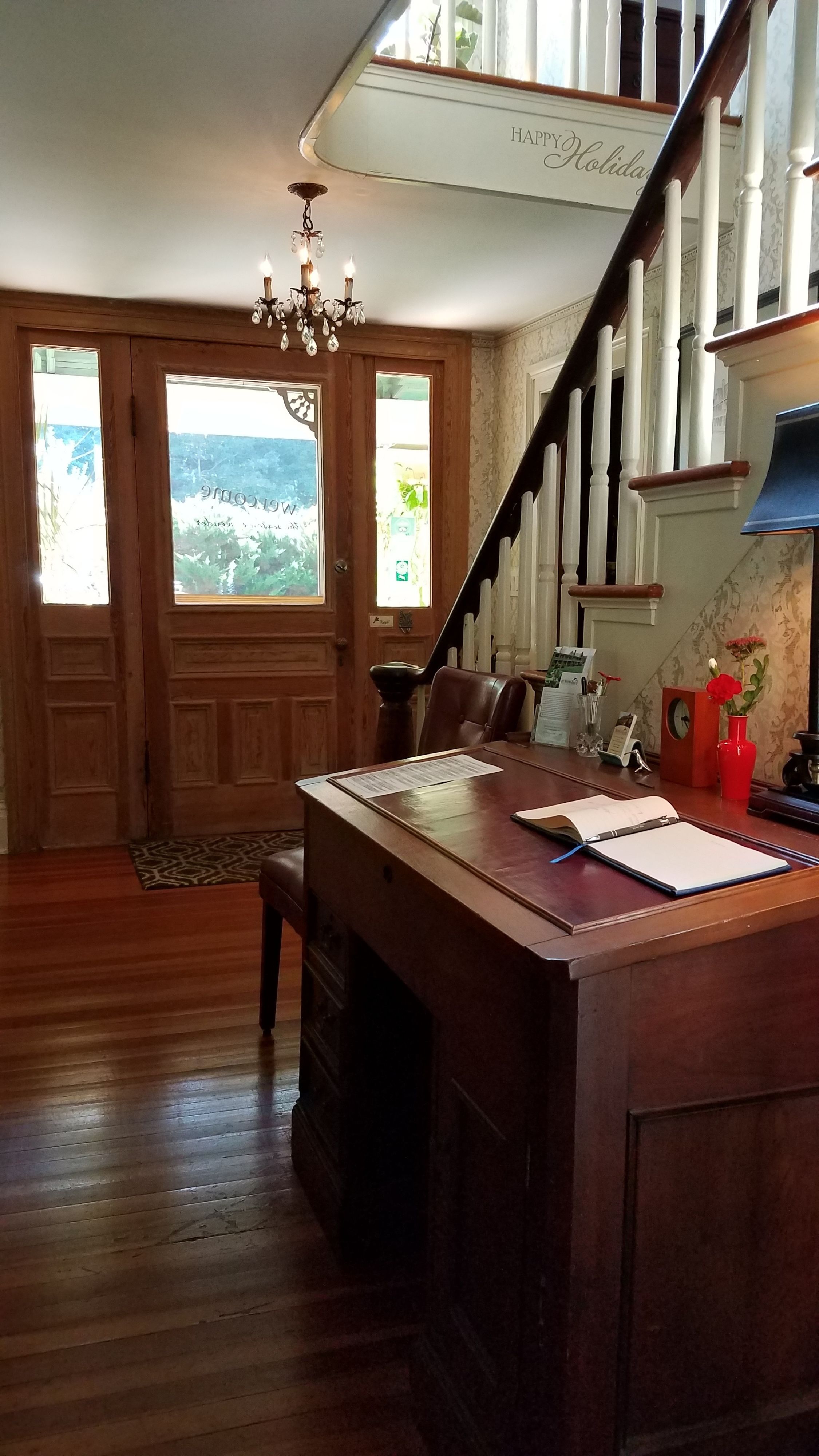 A spacious room with a wooden floor, a large wooden dining table, a chandelier, and a staircase with a 'Happy Holiday' sign.