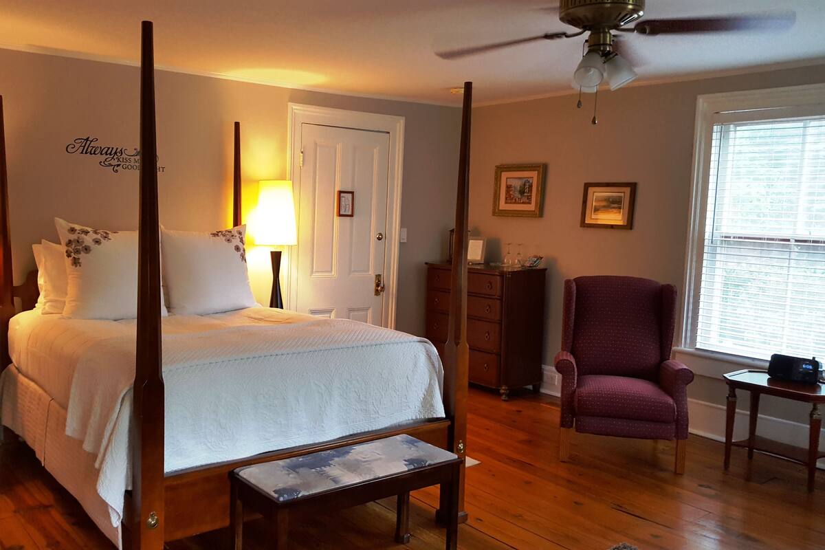 A four-poster bed with a white bedspread and floral pillows, a wooden nightstand with a lamp, a wooden bench, a purple armchair, and a wooden dresser with a mirror.