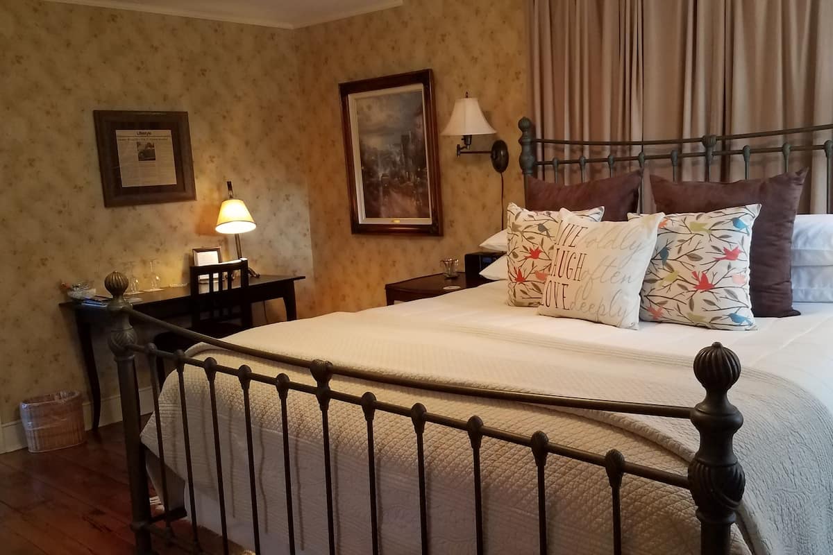 A neatly made bed with a metal frame, a bedside table with a lamp, a framed picture on the wall, and a window with curtains.