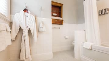 Un baño con piso de baldosas blancas, un armario de madera para medicinas, una bañera blanca y una bata blanca colgada en un perchero.