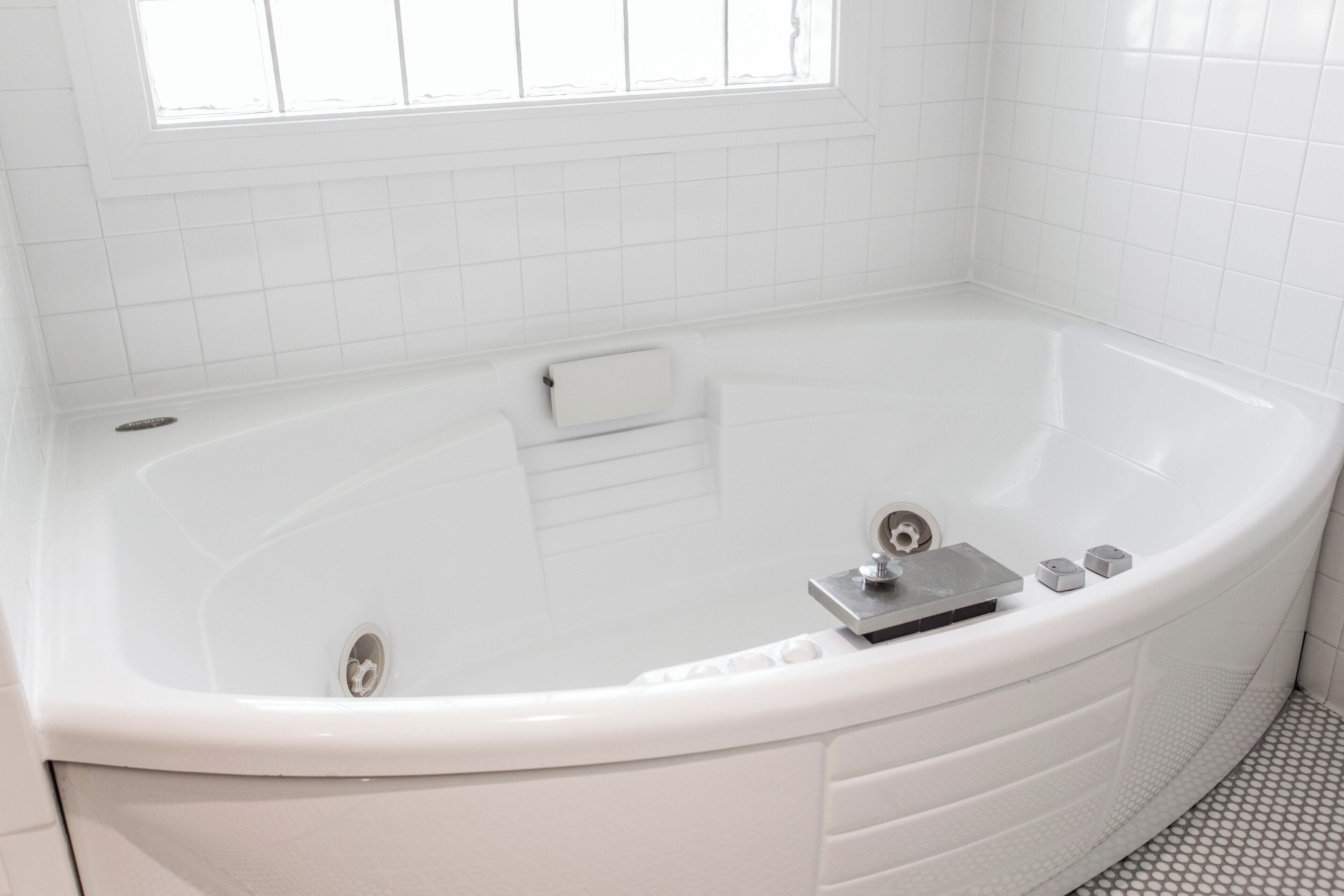 A white bathtub with a control panel, a window above it, and a tiled floor.