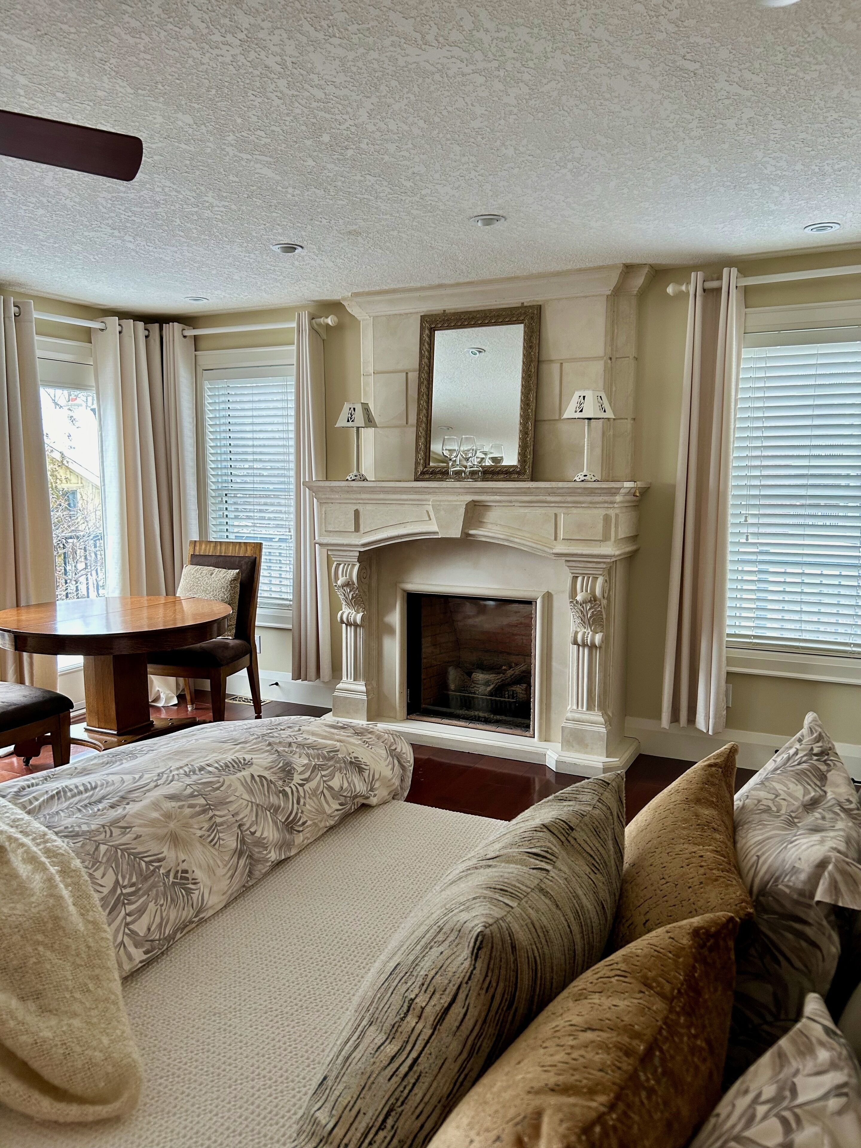 A room with a fireplace, a dining table with chairs, a bed with multiple pillows, and a window with blinds.