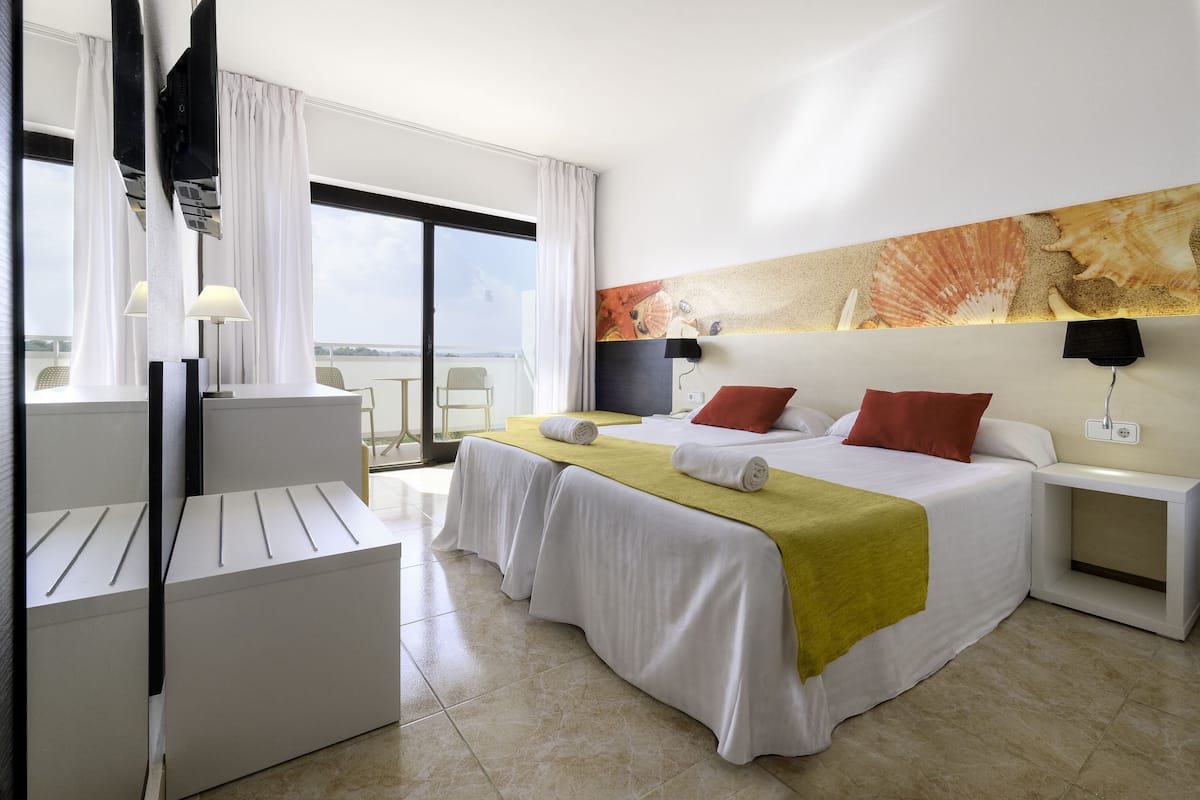 Ein Hotelzimmer mit Bett, Schreibtisch und Stuhl. Ein großes Fenster mit Vorhängen, ein Gemälde an der Wand und Blick aufs Meer.