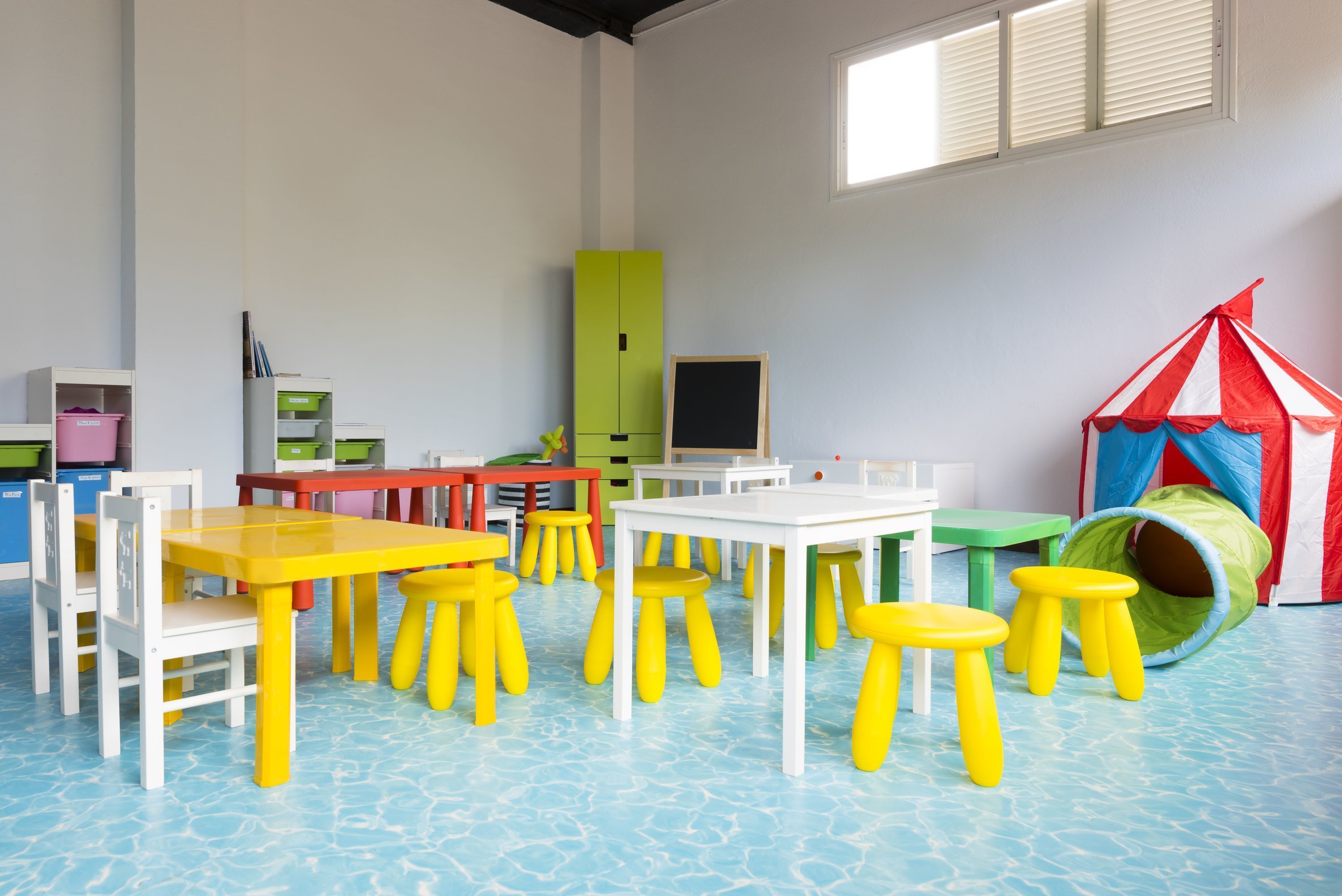 A brightly colored playroom with yellow and white tables and chairs, a green cabinet, and a red and white play tent.