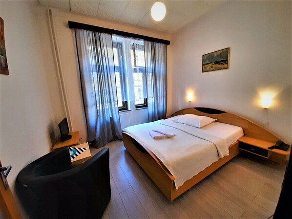 A hotel room with a bed, a desk, a chair, a window with curtains, and a painting on the wall.