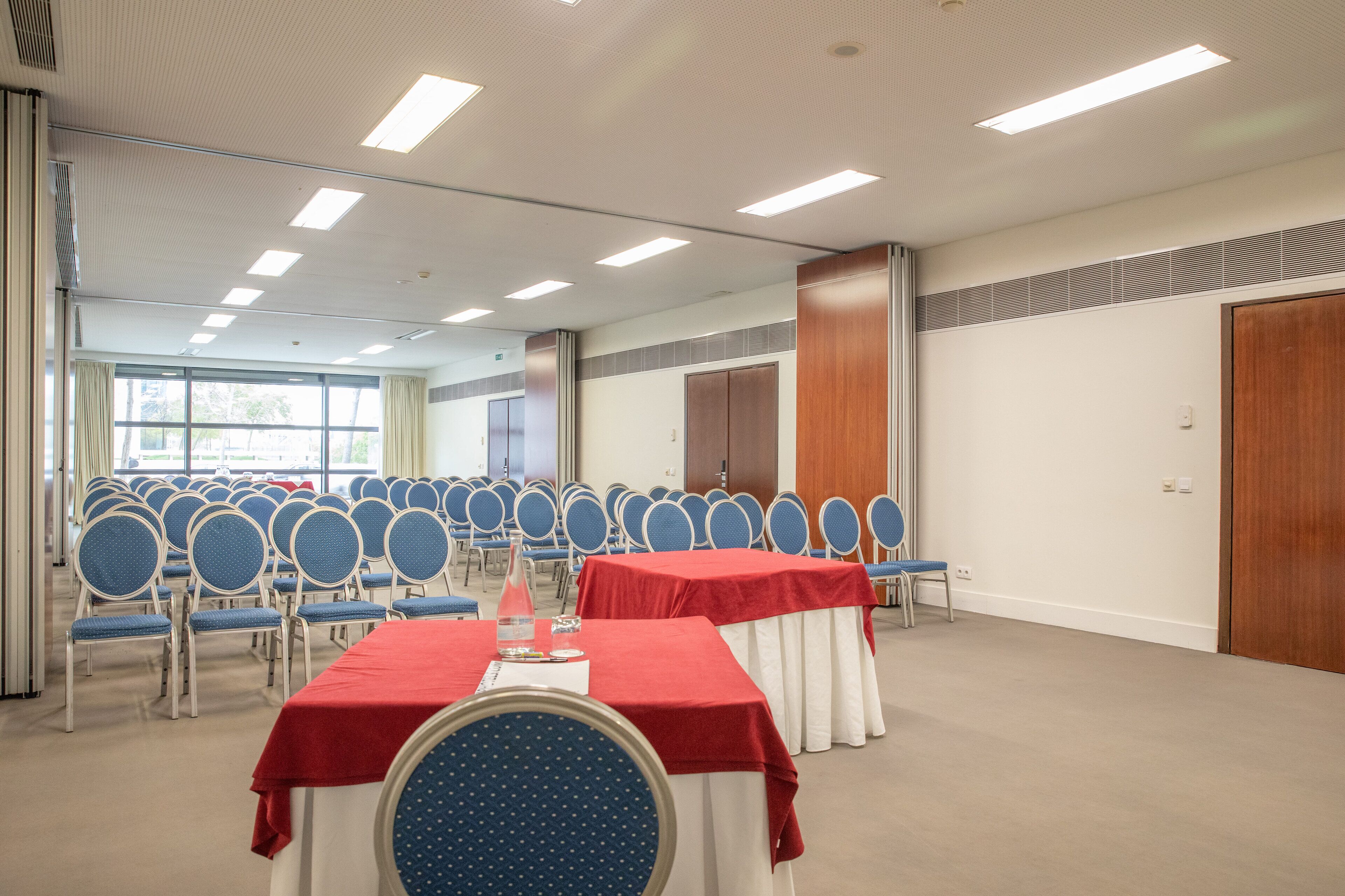 A conference room with rows of blue chairs, tables with red and white tablecloths, and a large window with curtains.