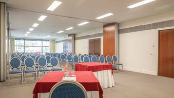 A conference room with rows of blue chairs, tables with red and white tablecloths, and a large window with curtains.