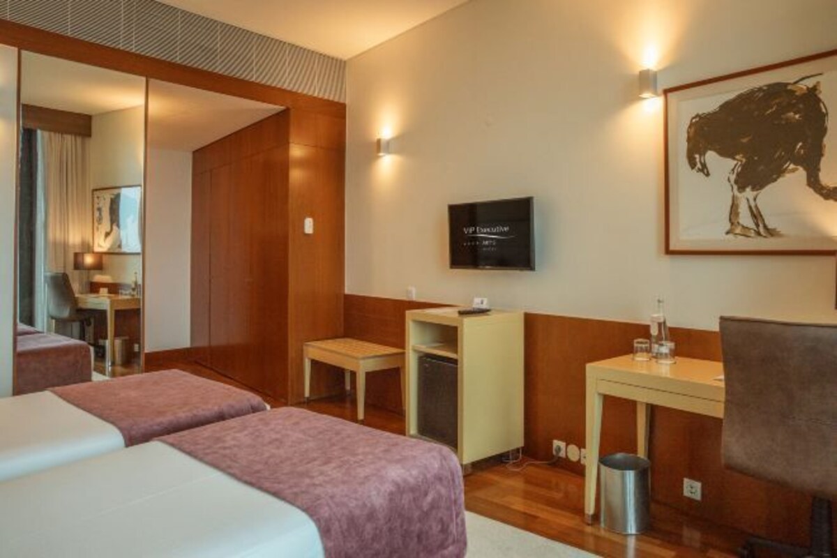 Una habitación de hotel con dos camas, un escritorio, una silla, un televisor y una obra de arte enmarcada en la pared.