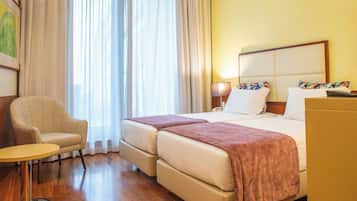 Habitación de hotel con cama, dos mesitas de noche, una silla, una mesa redonda pequeña y un ventanal grande con cortinas.