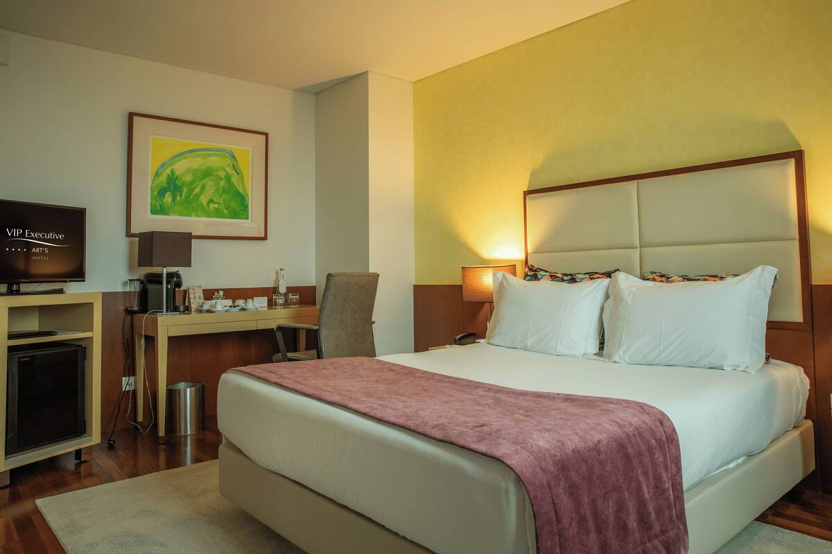 A hotel room with a bed, a desk, a television, and a painting on the wall.