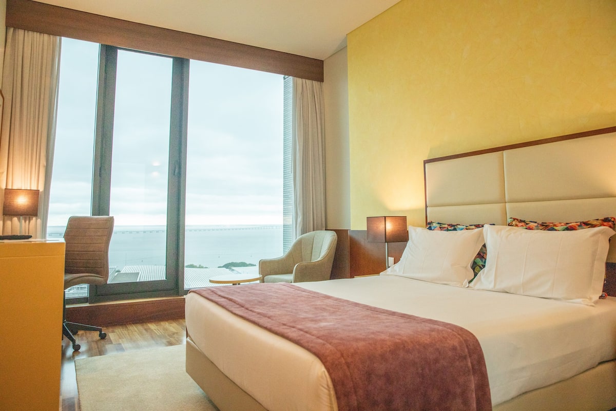 A hotel room with a large bed, two bedside tables, a desk, and a chair. There are curtains on the windows, and a view of the sea is visible.