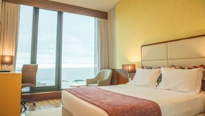 Habitación de hotel con una cama grande, dos mesitas de noche, un escritorio y una silla. Hay cortinas en las ventanas y se ve el mar.