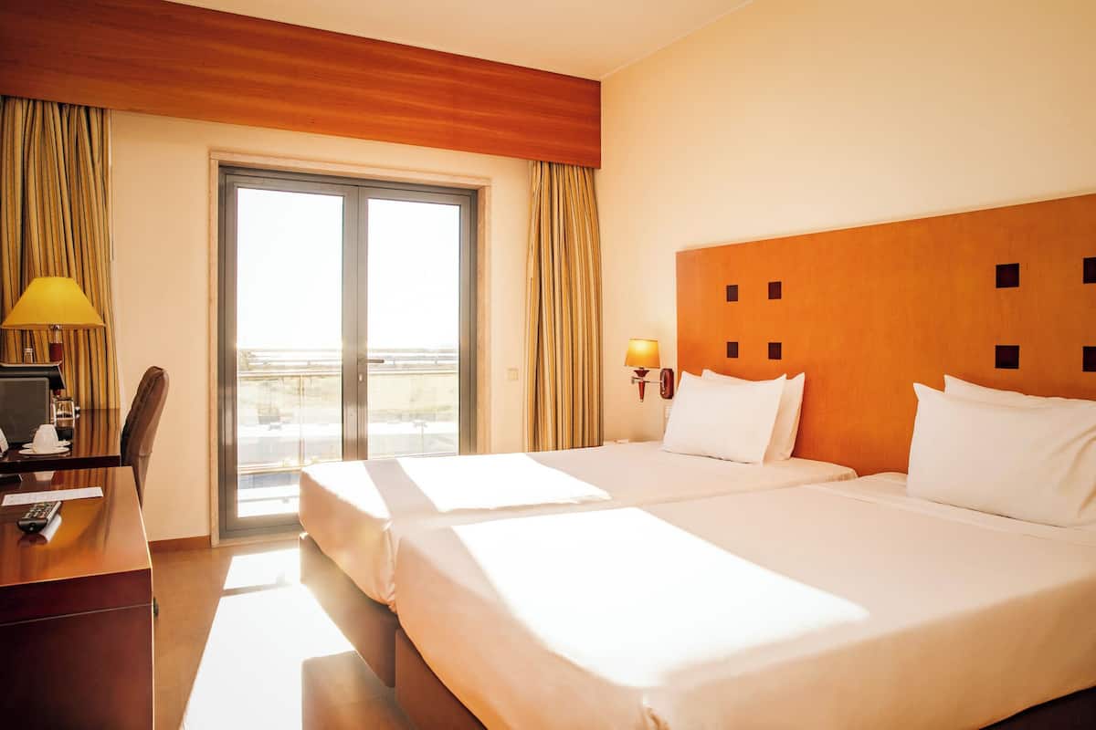 Ein Hotelzimmer mit zwei Betten, einem Schreibtisch mit Computer, einer Lampe und Blick ins Freie.