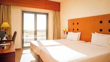 Habitación de hotel con dos camas, un escritorio con lámpara, una silla y vistas al exterior a través de una puerta corrediza.