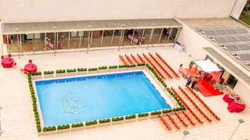 A swimming pool with a dolphin design, surrounded by red chairs and greenery, with a covered area and solar panels in the background.