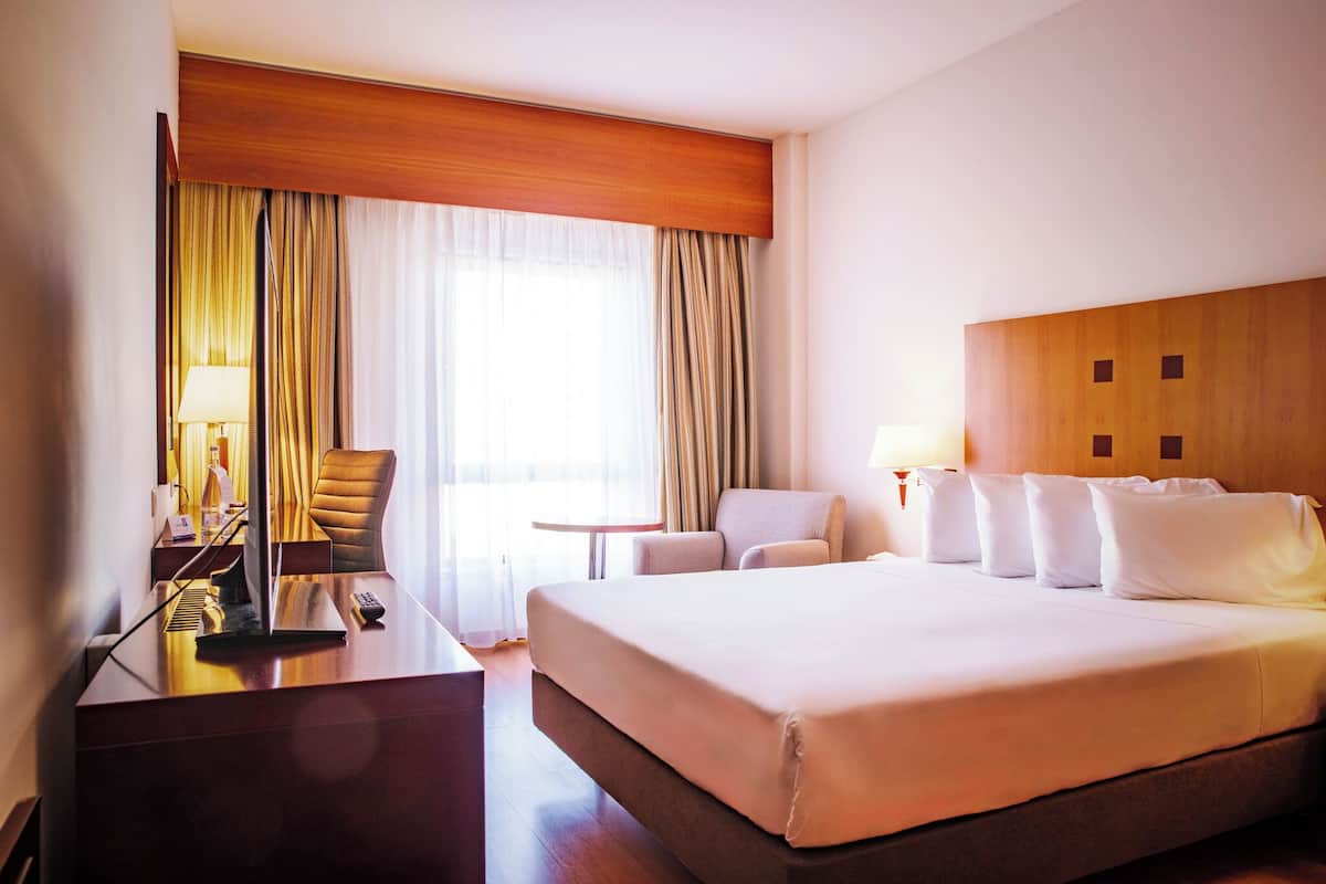 Ein Hotelzimmer mit einem großen Bett, einem Schreibtisch mit Computer, einem Stuhl, einem kleinen Tisch und einem Fenster mit Vorhängen.