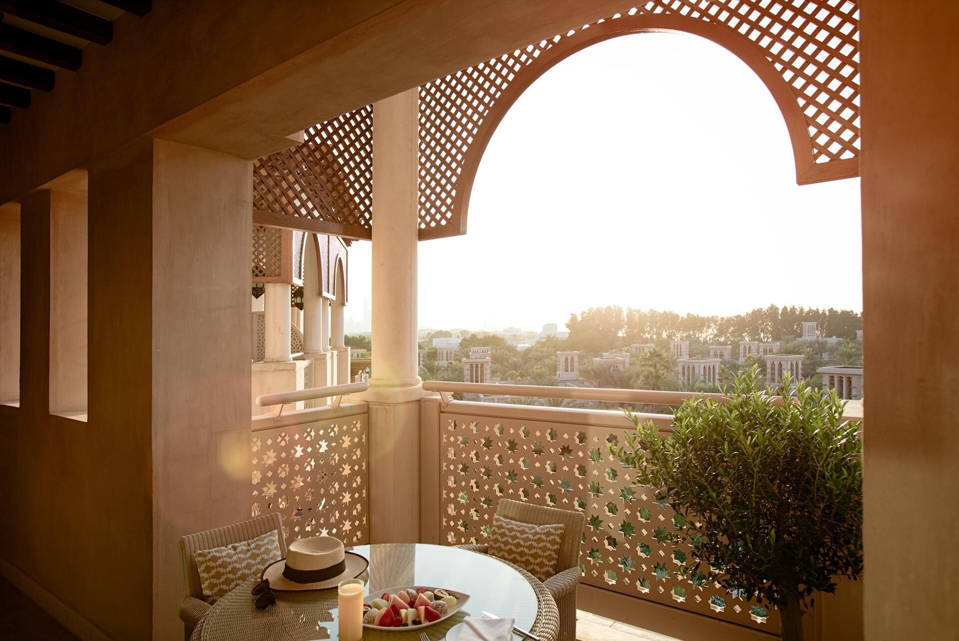 A balcony with a view of a cityscape, featuring a wicker chair, a small table with a plate of fruit, and a decorative wall with star-shaped cutouts.