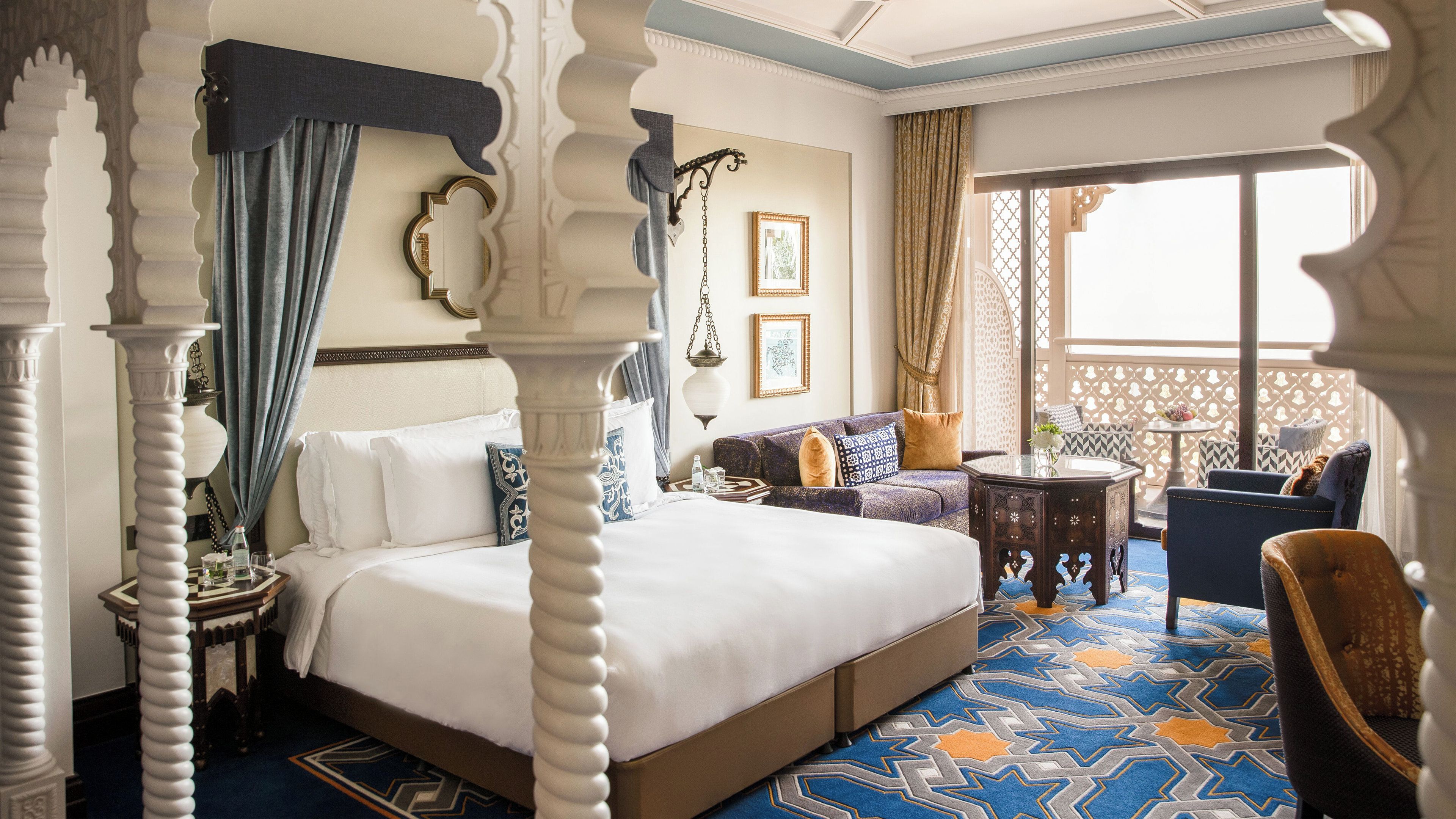 A four-poster bed with a canopy, a sofa with patterned cushions, a small round table, and a chair with a patterned cushion.