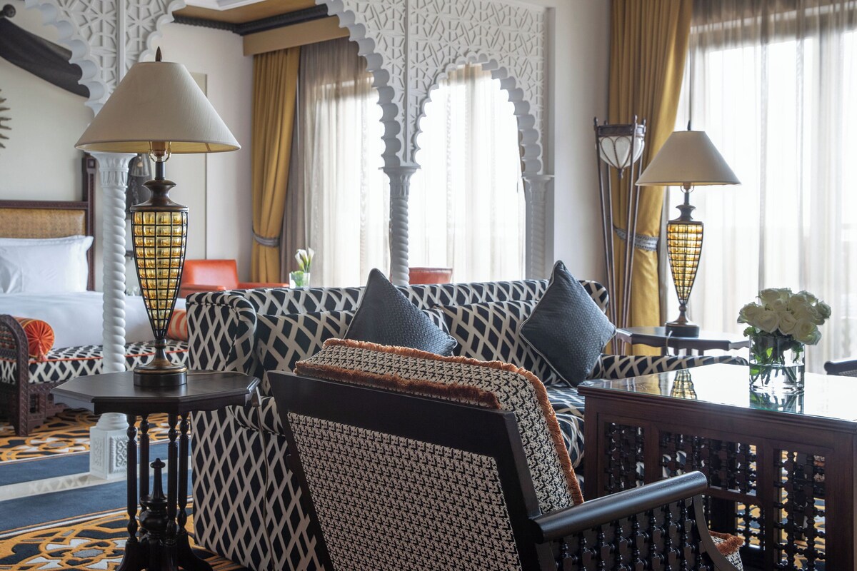 A hotel room with a bed, a sofa, two lamps, a vase with flowers, and a patterned carpet.