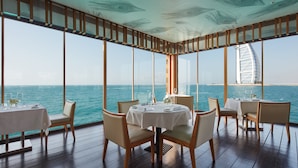 A dining area with tables set for four, large windows offering a view of the sea and a distinctive building in the background.