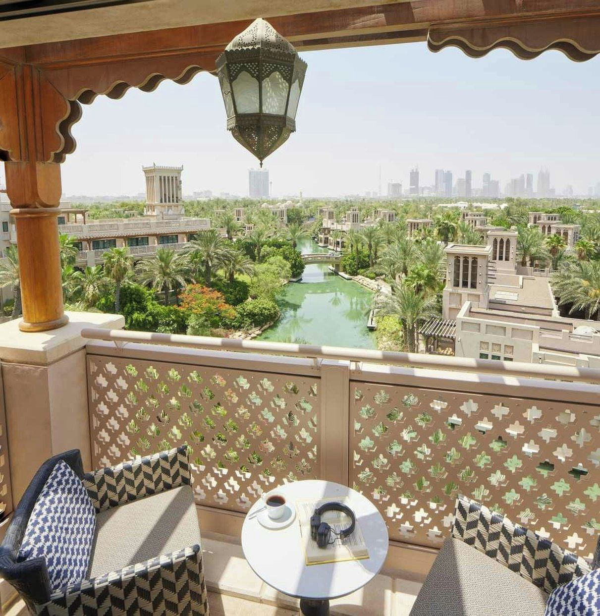 A balcony with a view of a water feature and buildings, a table with a cup and headphones, and patterned cushions.