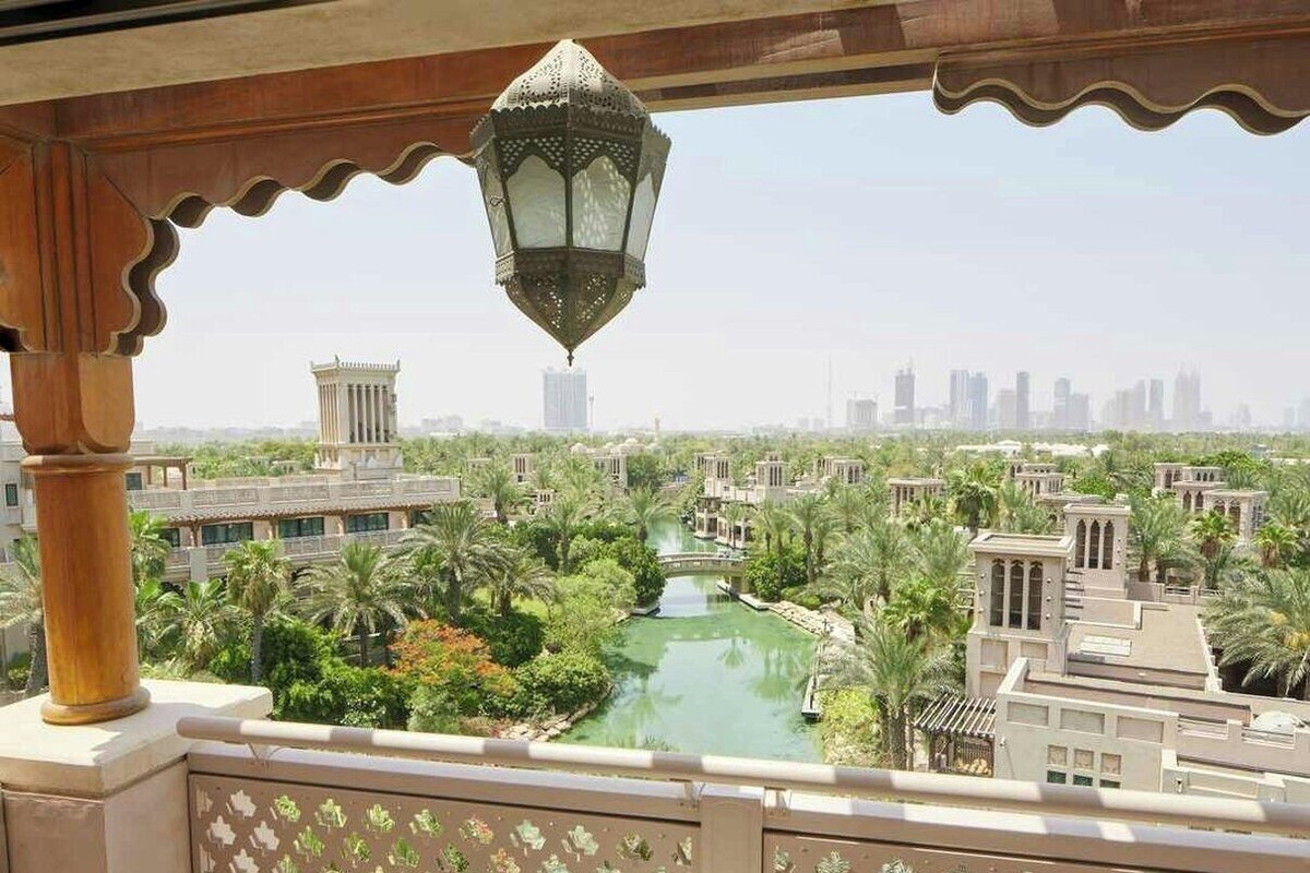 A balcony with a view of a water feature and buildings, a table with a cup and headphones, and patterned cushions.