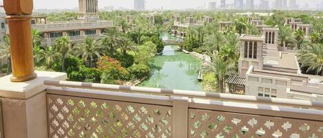 A balcony with a view of a water feature and buildings, a table with a cup and headphones, and patterned cushions.