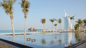 A pool area with palm trees, a bench, and a view of the sea and a distinctive building in the background. - Jumeirah Al Qasr Dubai (Dubai)