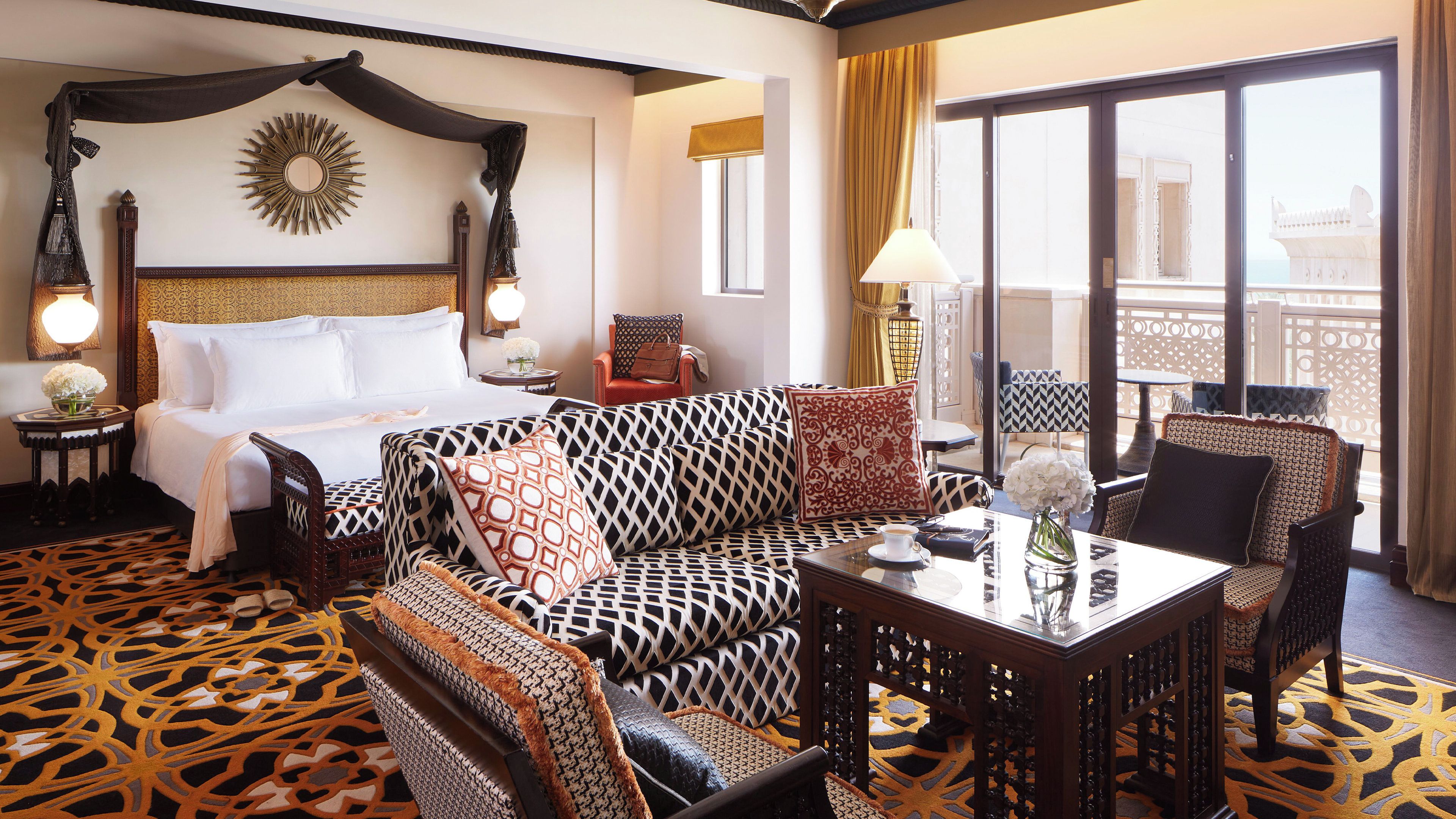 Habitación de hotel con una cama grande, un sofá estampado, una mesa de comedor con sillas y un balcón con vistas.