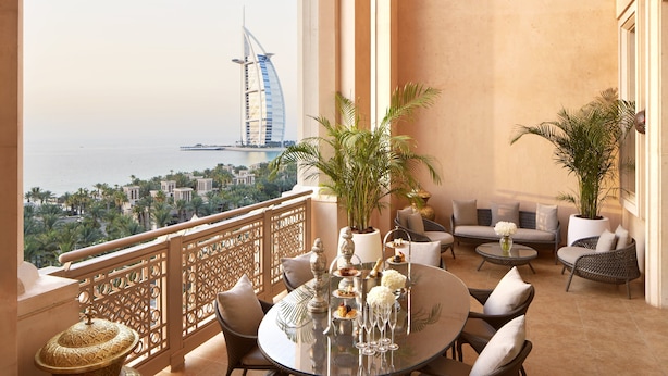 A balcony with a view of a sail-shaped building, furnished with wicker chairs and a glass table set for dining.