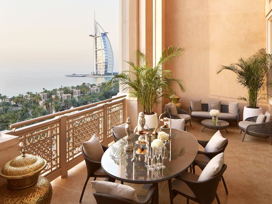 A balcony with a view of a sail-shaped building, furnished with wicker chairs and a glass table set for dining.