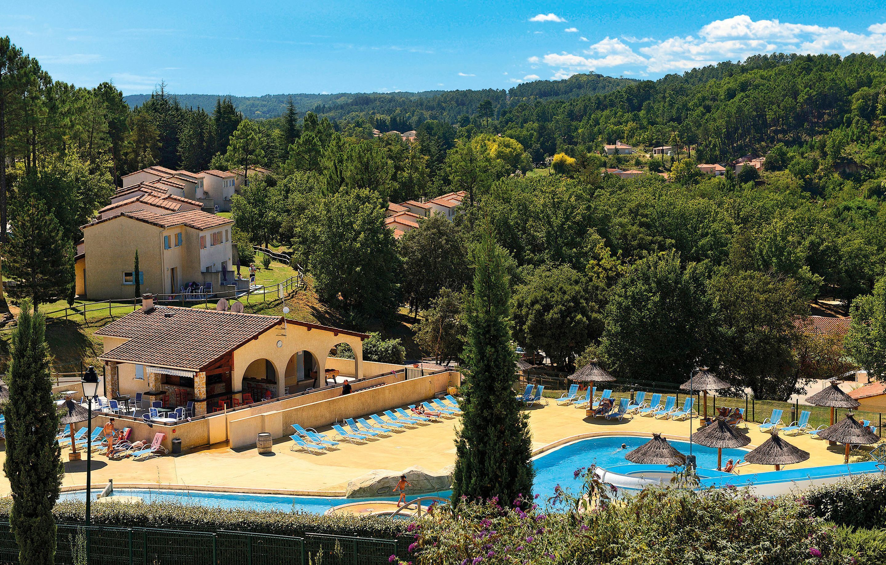 A resort with a swimming pool, sun loungers, and a view of the surrounding forest.