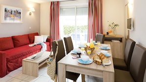 A modern living room with a red sofa, a dining table set with breakfast, and a large window with curtains. - Résidence Odalys Domaine des Hauts de Salavas (Salavas)