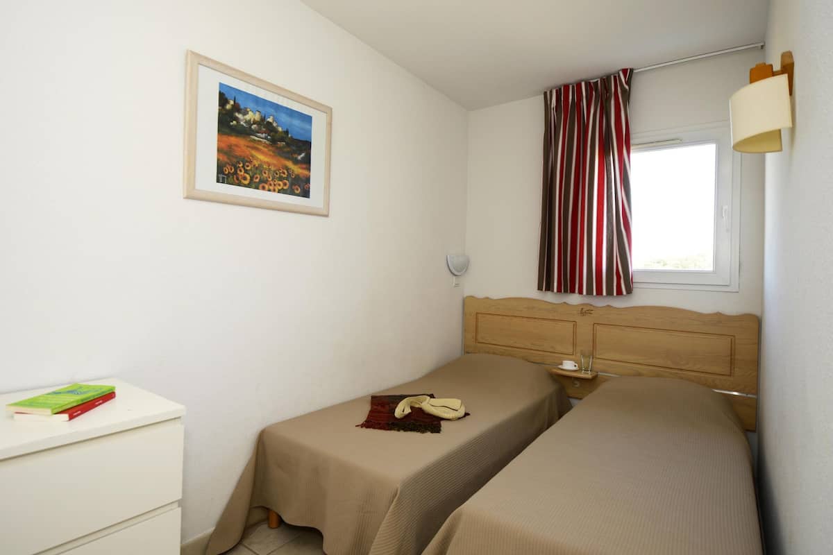Ein Schlafzimmer mit zwei Betten, einem Nachttisch mit Lampe, einem gerahmten Bild an der Wand und einem Fenster mit Vorhängen.