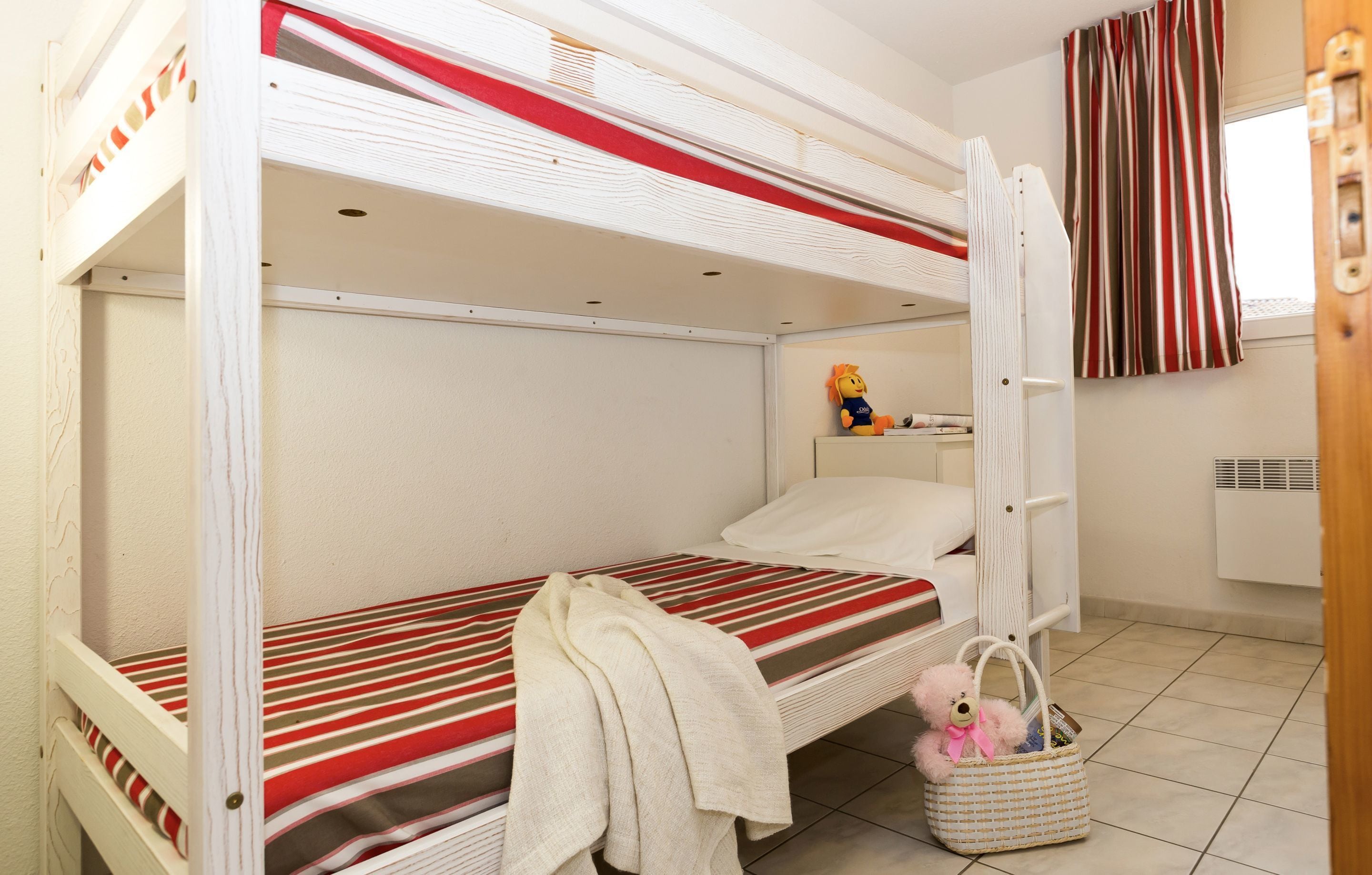 Une chambre avec un lit superposé, recouverte d’une couette à rayures rouges et blanches, une serviette blanche et un ours en peluche sur le lit du bas.