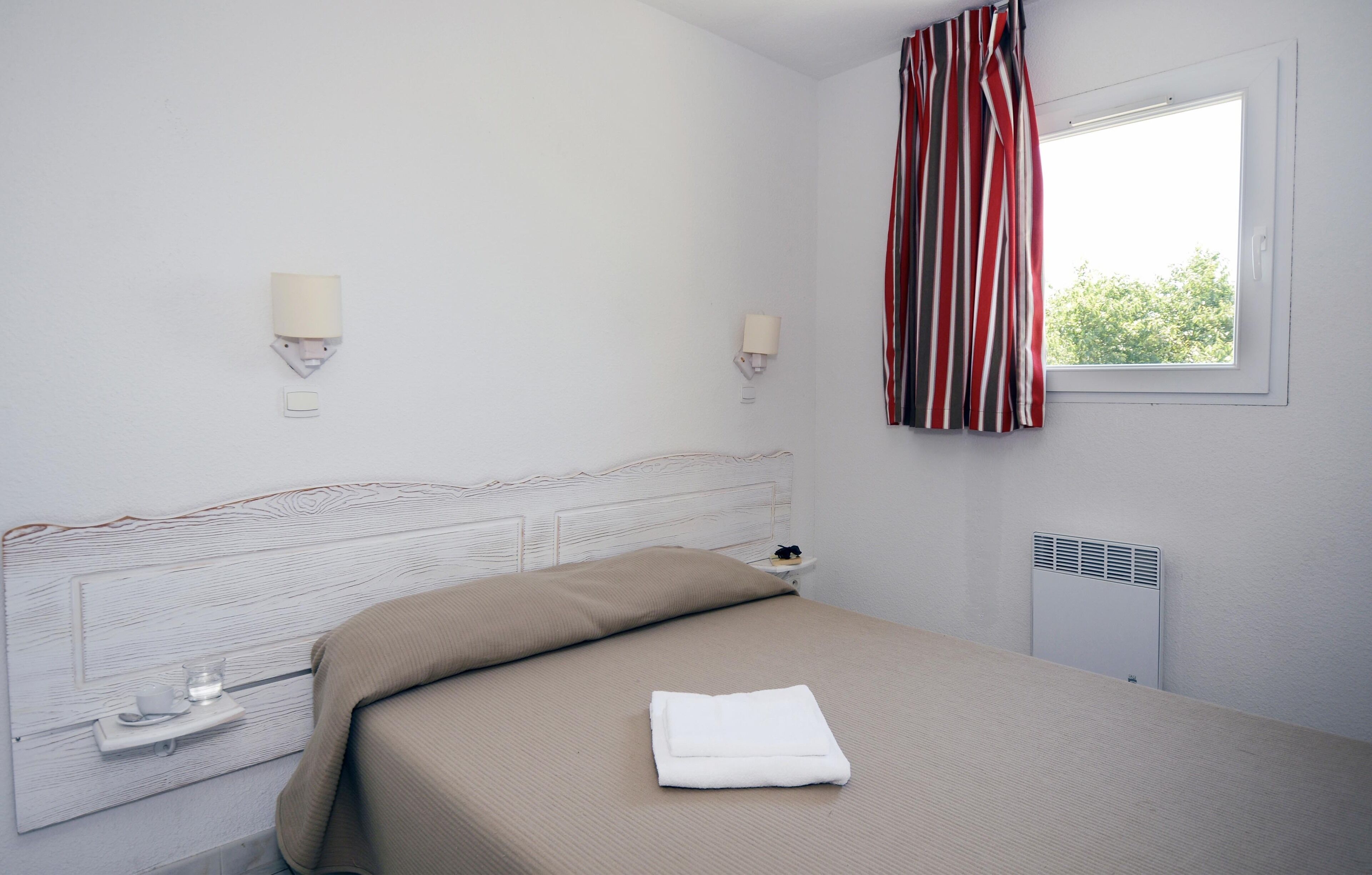 A neatly made bed with a folded towel, a bedside table with a glass, and a window with curtains.