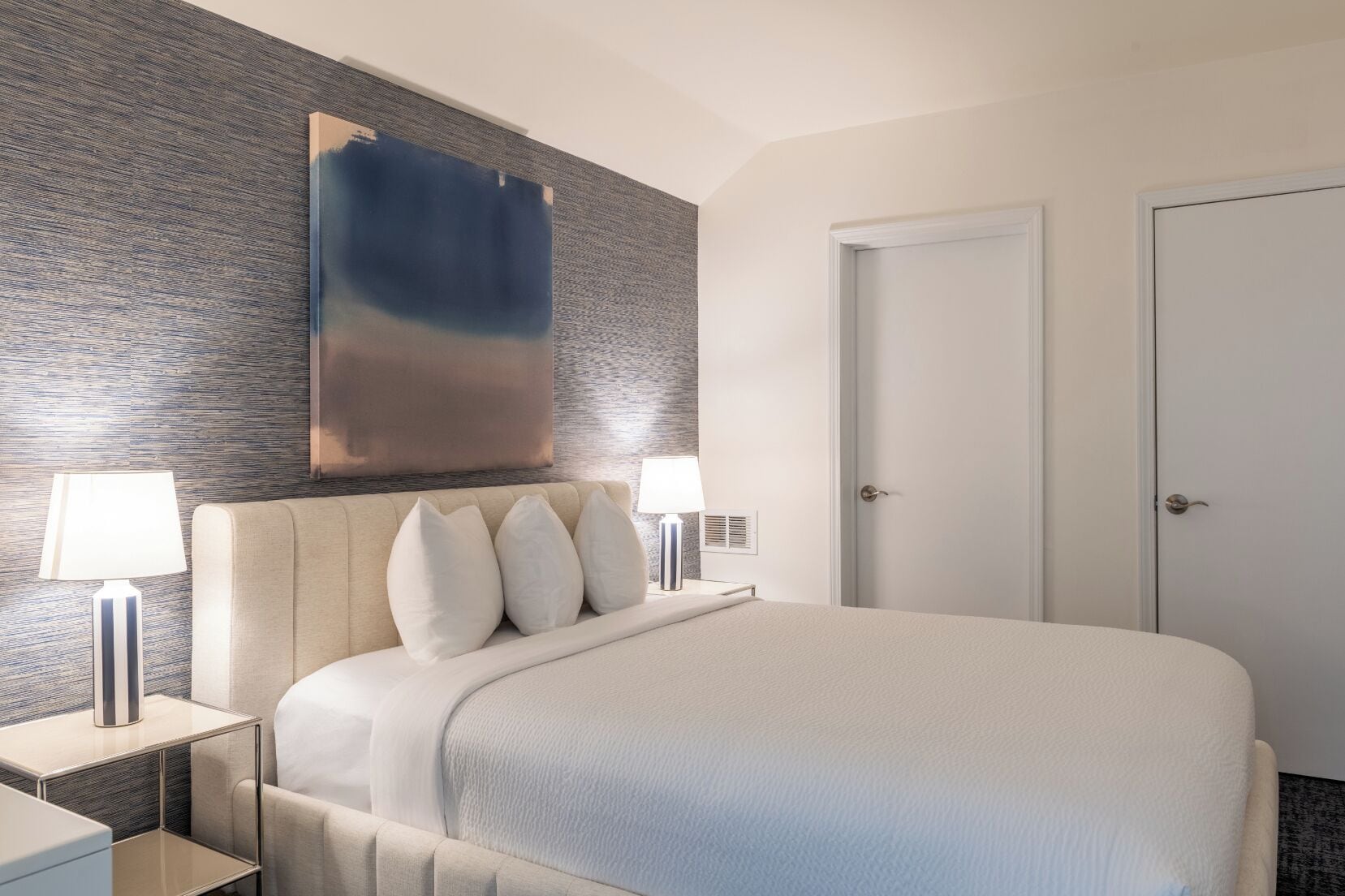 A hotel room with a large bed, two bedside tables with lamps, a modern abstract painting on the wall, and a door.