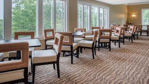 A dining area with multiple tables and chairs, large windows with a view of trees, and a patterned carpet.