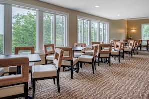 A dining area with multiple tables and chairs, large windows with a view of trees, and a patterned carpet.