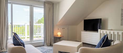 A modern living room with a white sofa, a coffee table, and a TV mounted on the wall. There is a balcony with a view of greenery outside.