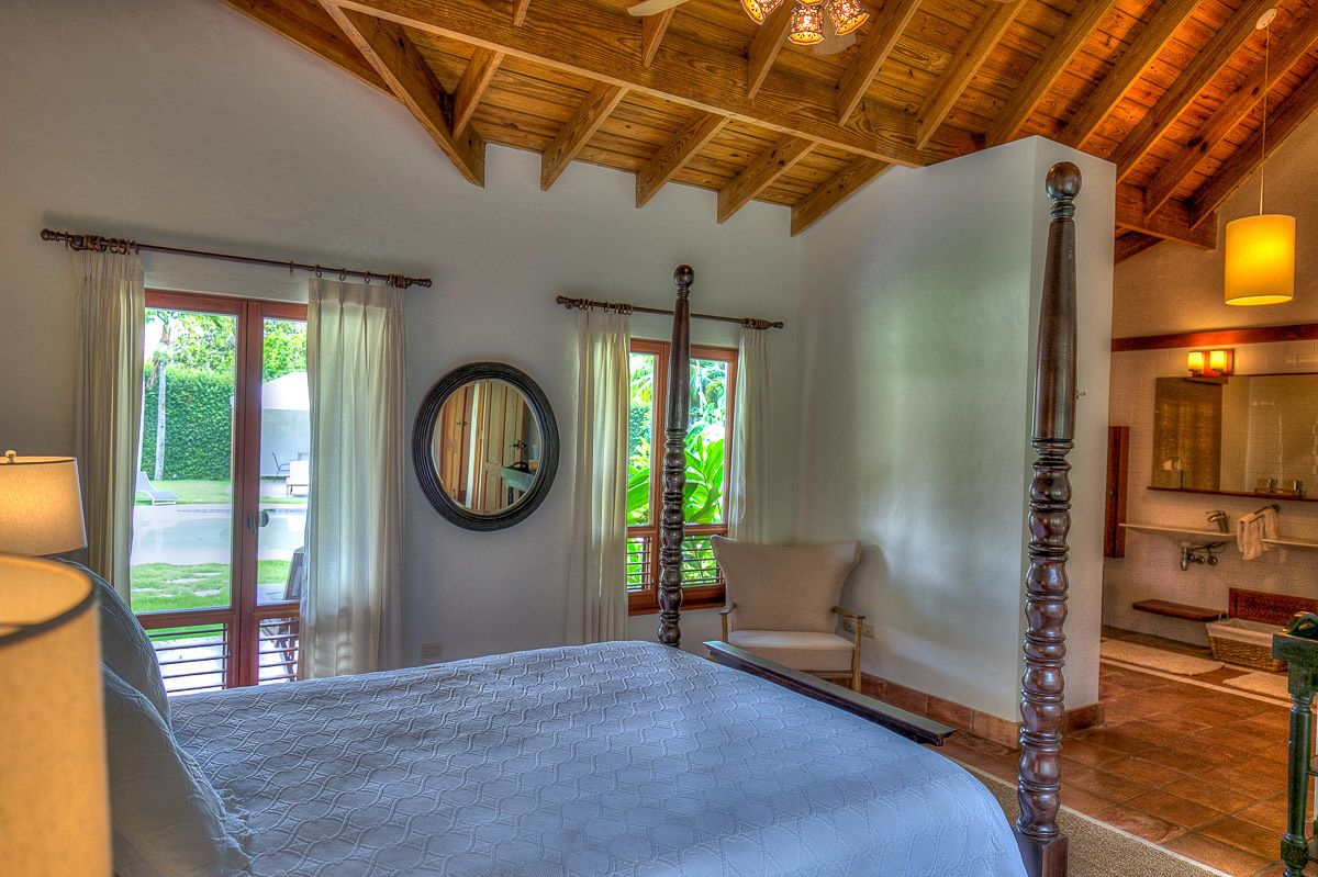 A bedroom with a large bed, wooden ceiling beams, a mirror, and a view of the garden through the window.