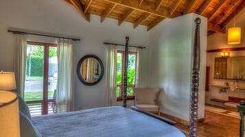 A bedroom with a large bed, wooden ceiling beams, a mirror, and a view of the garden through the window.