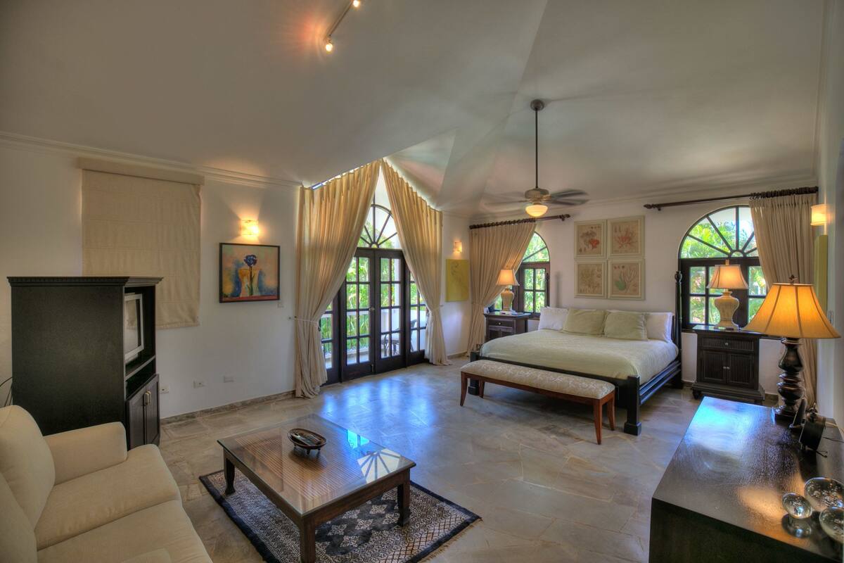 A spacious bedroom with a large bed, a desk, and a sofa. There are multiple windows with curtains, a ceiling fan, and artwork on the walls.