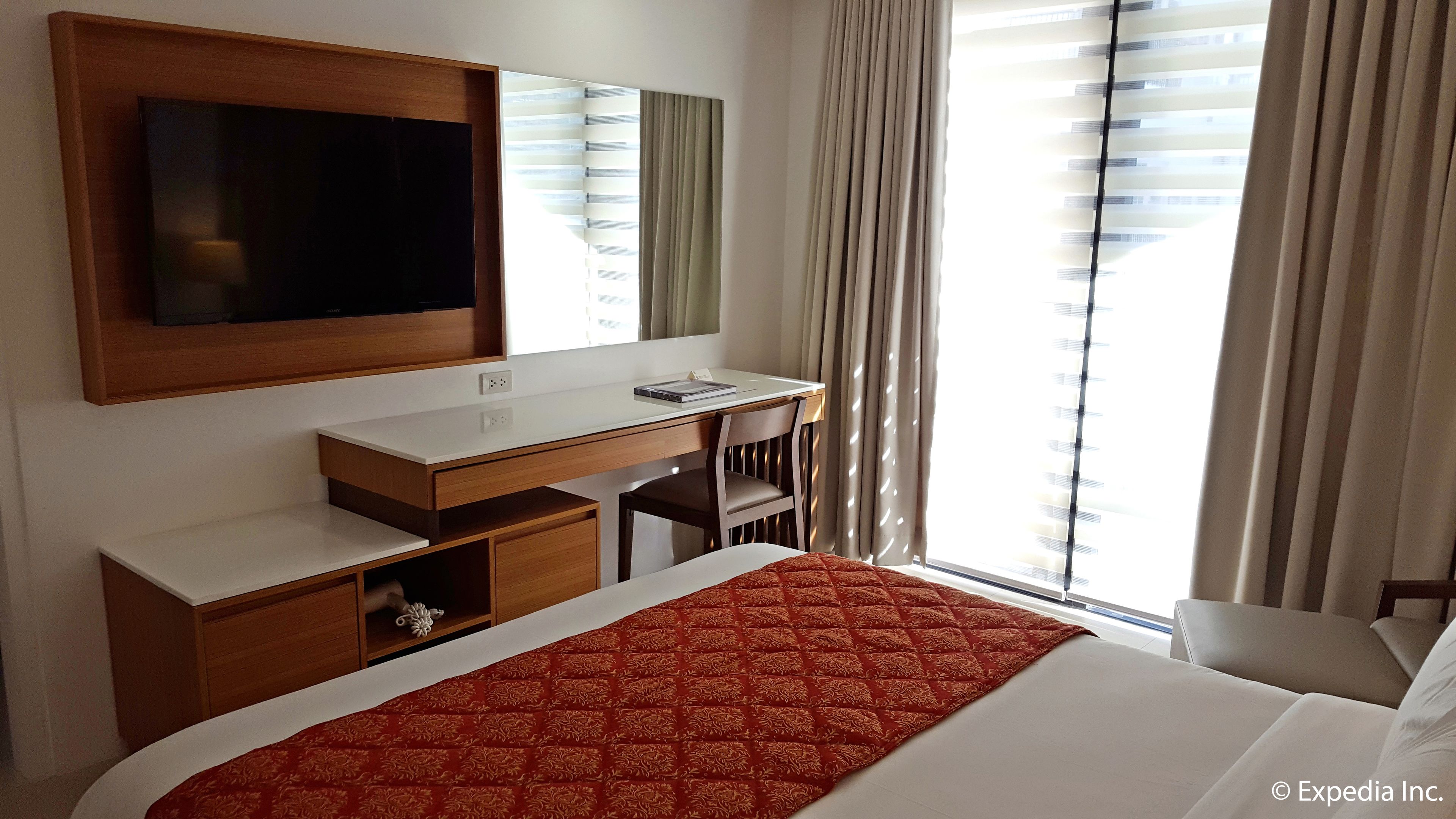 Habitación de hotel con cama, escritorio, silla, televisor y ventana con cortinas.
