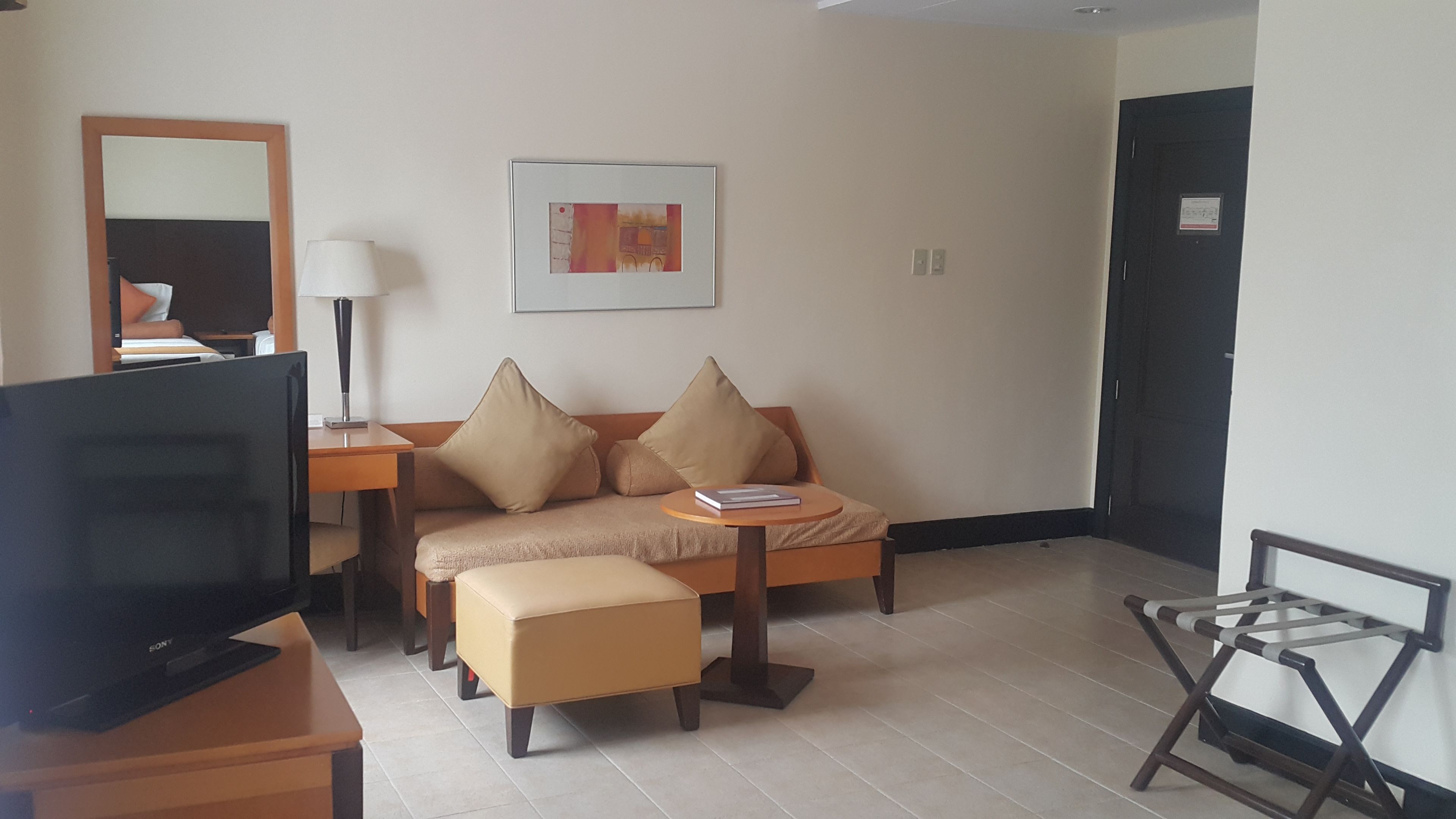 A hotel room with a sofa, ottoman, TV, lamp, and a framed picture on the wall.