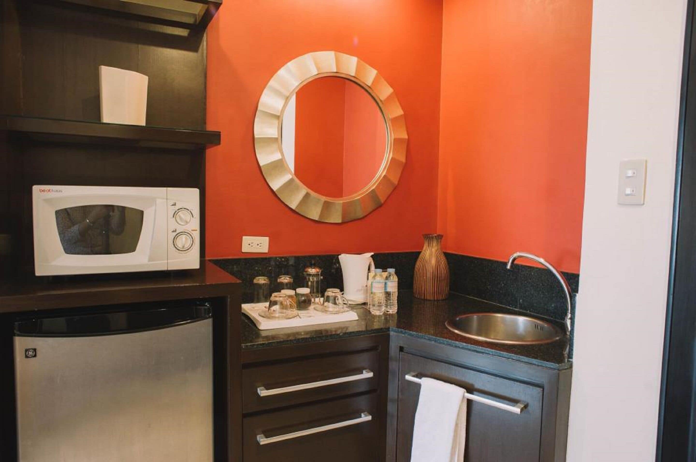 A compact kitchen with a microwave, refrigerator, sink, and a decorative mirror.