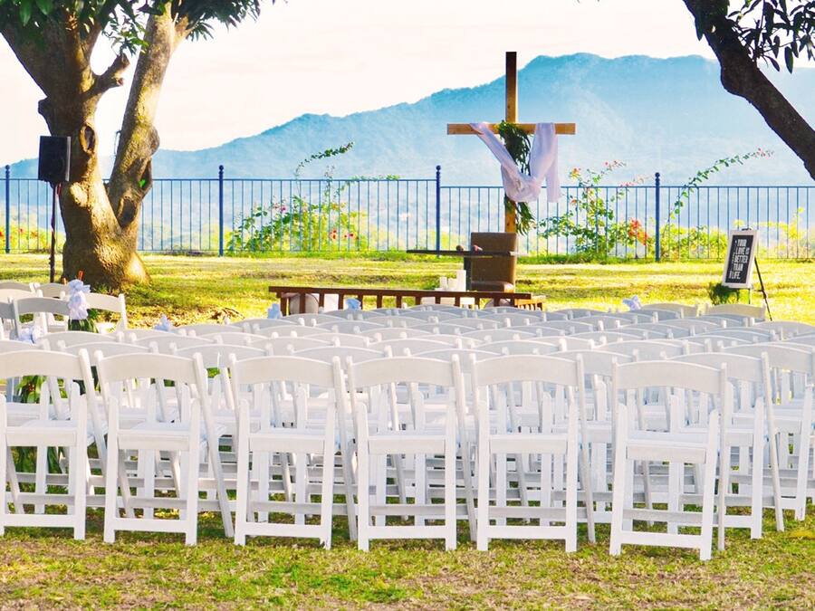 Montaje de ceremonia de boda con sillas blancas dispuestas en filas, un banco de madera y una cruz.